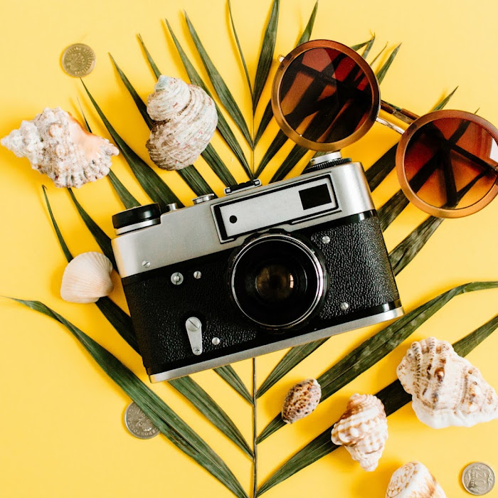 Flat lay of a vintage camera and sunglasses on palm leaves with seashells and coins, against a bright yellow background
