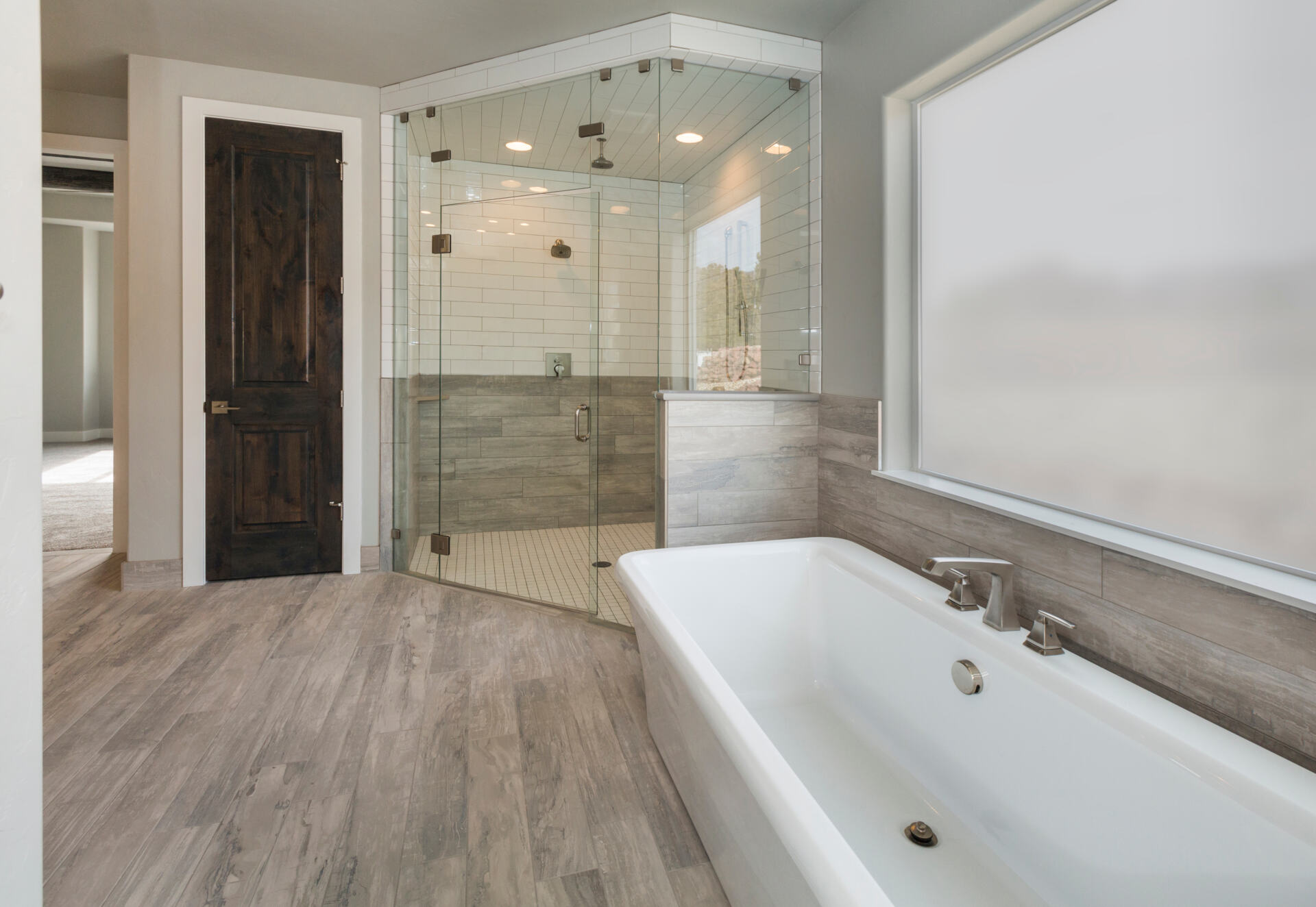 Modern bathroom with a freestanding white soaking tub, glass-enclosed tiled shower, wood-look tile flooring, and a dark accent wall