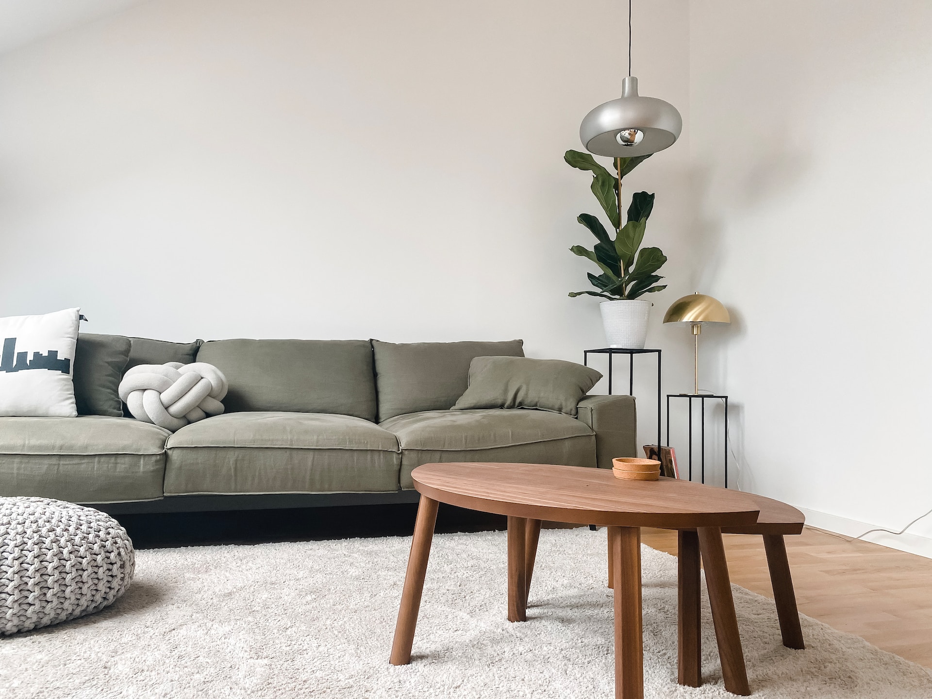 A cozy living room with an olive-green sofa, knotted cushion, round wooden coffee table, knit pouf, pendant and floor lamp